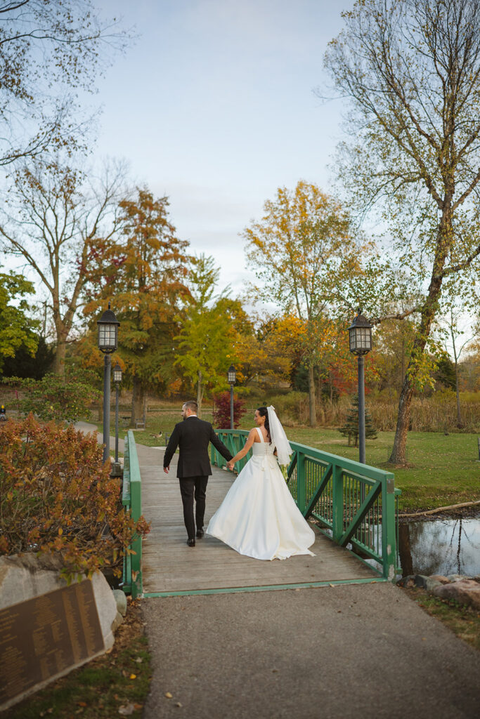Bride and groom walking together across a bridge in fall during portraits at a Clarkston Michigan wedding.