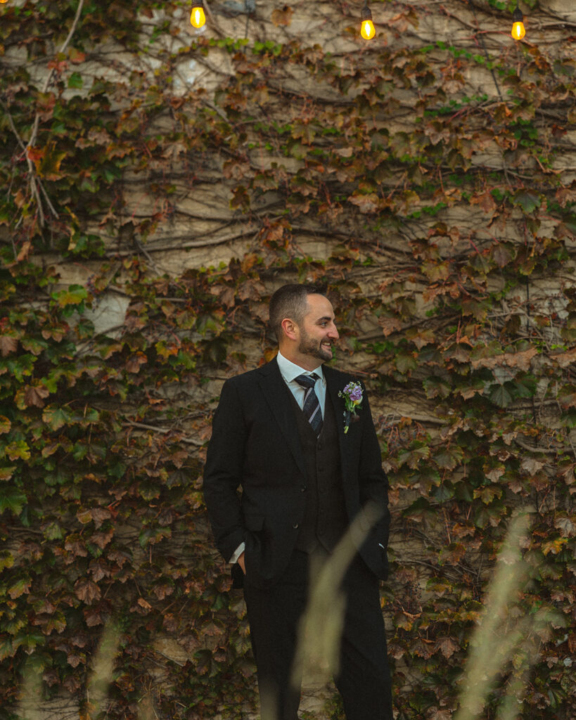 Outdoor portrait of the groom standing in front of an ivy covered wall. 