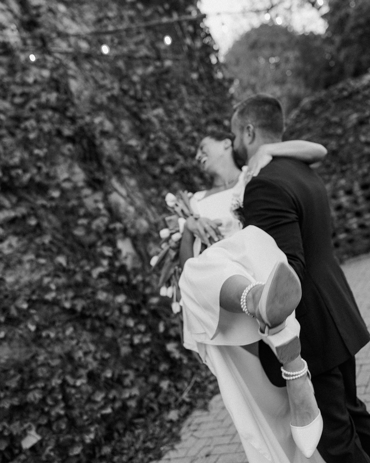 Black and white candid photo of a bride laughing while the groom lifts her in his arms. 