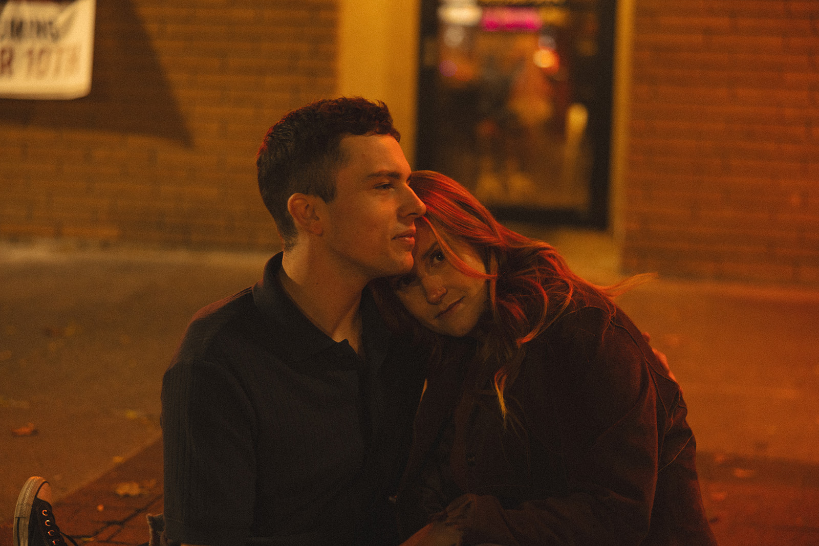 Close up shot of a woman leaning into her fiancé as they sit on a sidewalk. 