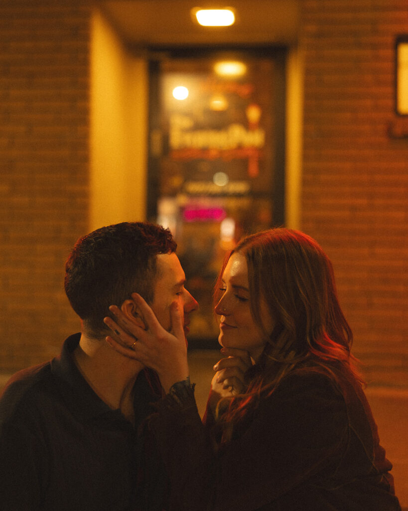 Woman caressing her fiancés face as they sit on a sidewalk outside of a dive-bar.