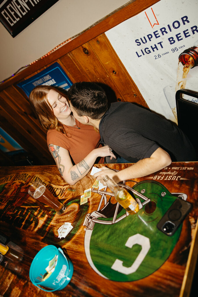 Flash photo of a couple laughing while having beers and playing pull tab at The Evening Post Bar & Grill in Clare, Michigan for their dive bar engagement photos.