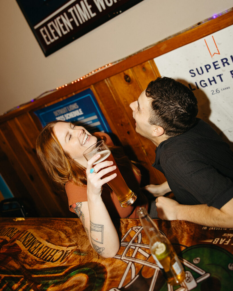 Candid flash photo of a couple laughing while having drinks during their dive bar engagement photos.
