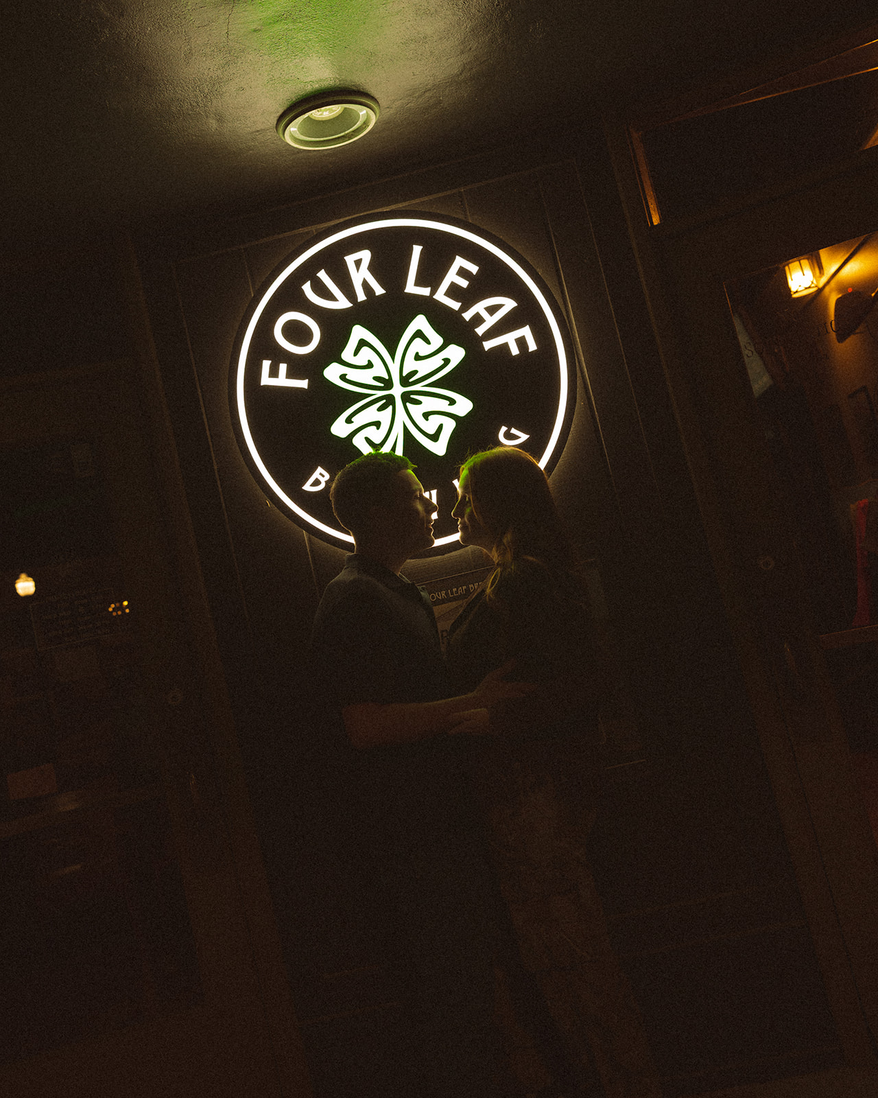 Dark photo of a couple holding on to one another in front of a sign that says Four Leaf Brewing. during their dive bar engagement photos.