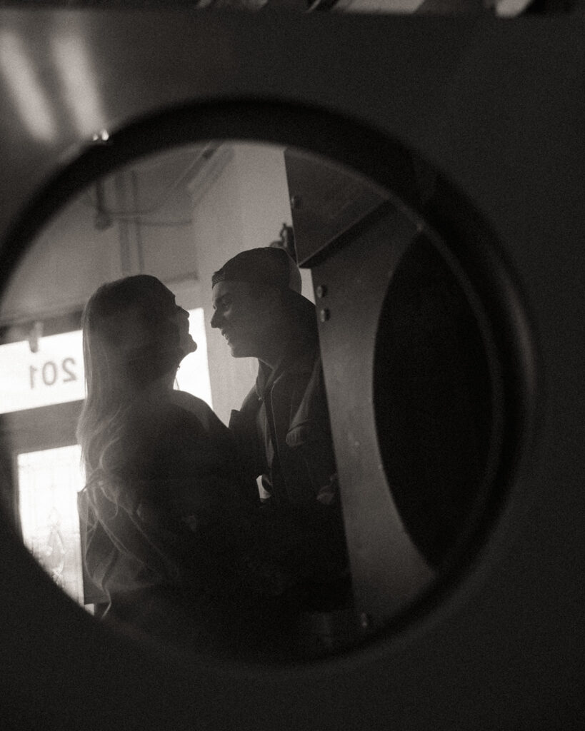 Black and white photo through a washer window of a couple being playful at their local laundromat.