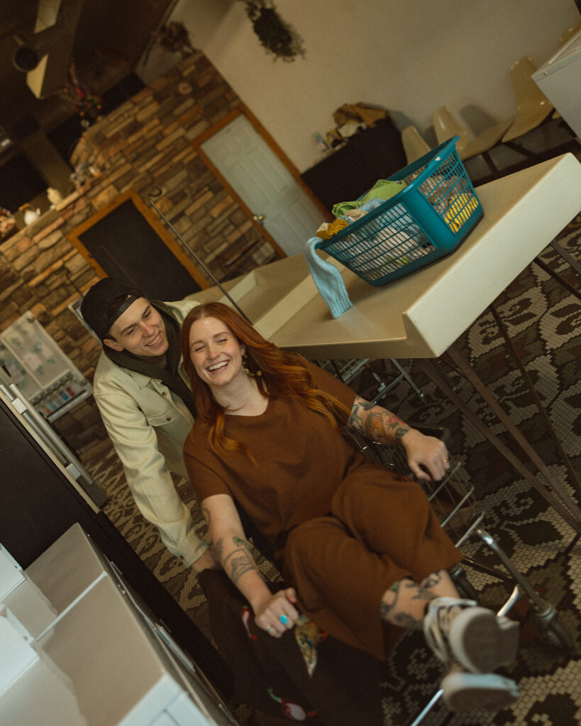 Couple laughing as a man pushes his fiancé in a laundromat basket during their photoshoot.