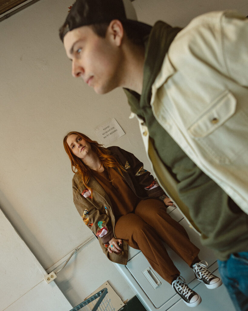 Woman watching her fiancé as he stands in a Michigan laundromat for their photoshoot.