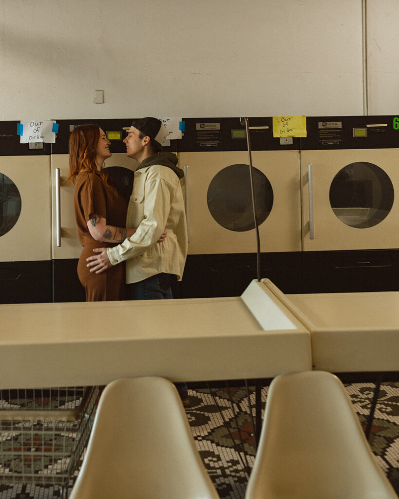 Couple holding on to one another in front of the washing machines during their laundromat photoshoot in Michigan.