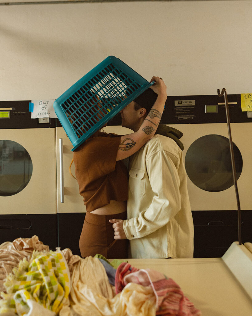 Couple posing with a laundry basket  over their heads during their cute laundromat photoshoot for their engagement.