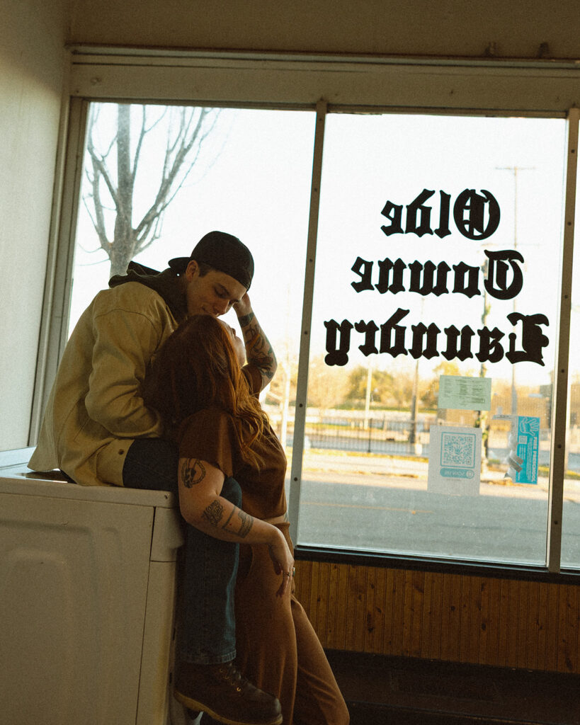 Couple posing in front of a washing machine and window for their laundromat photoshoot.