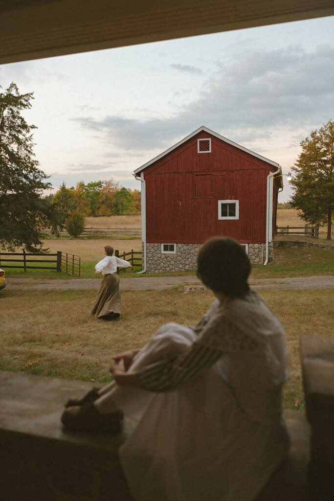 One partner sitting on the farmhouse porch while the other runs across the yard toward a red barn.