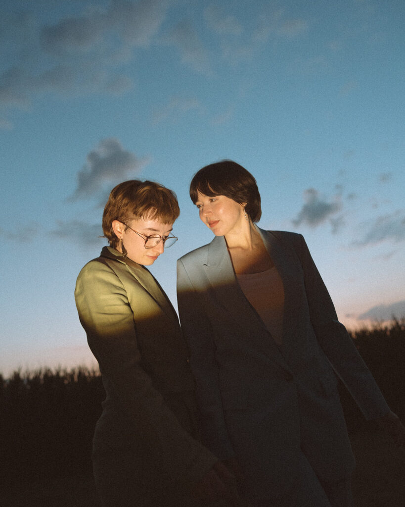 Couple in colorful suits looking at each other beneath a dusky blue sky and direct flash.