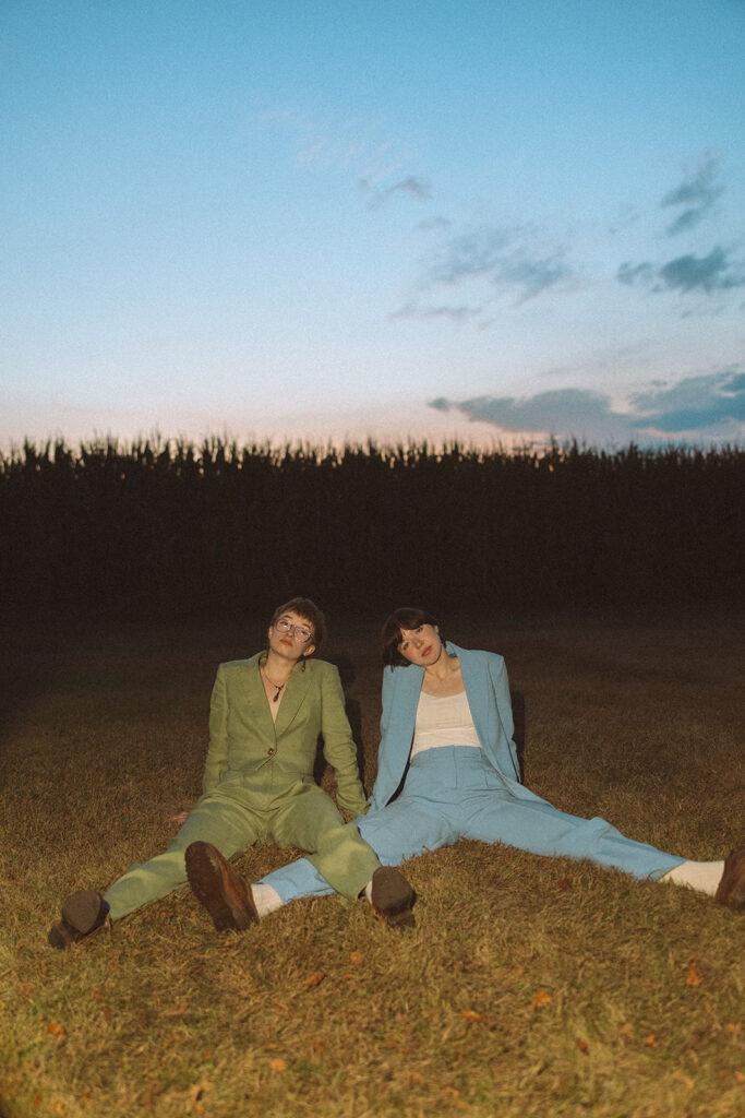 Couple sitting together in the grass beside a cornfield during their Midwestern Gothic engagement photos.