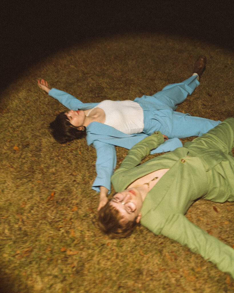 Couple lying side by side in the grass while wearing coordinating green and blue suits.