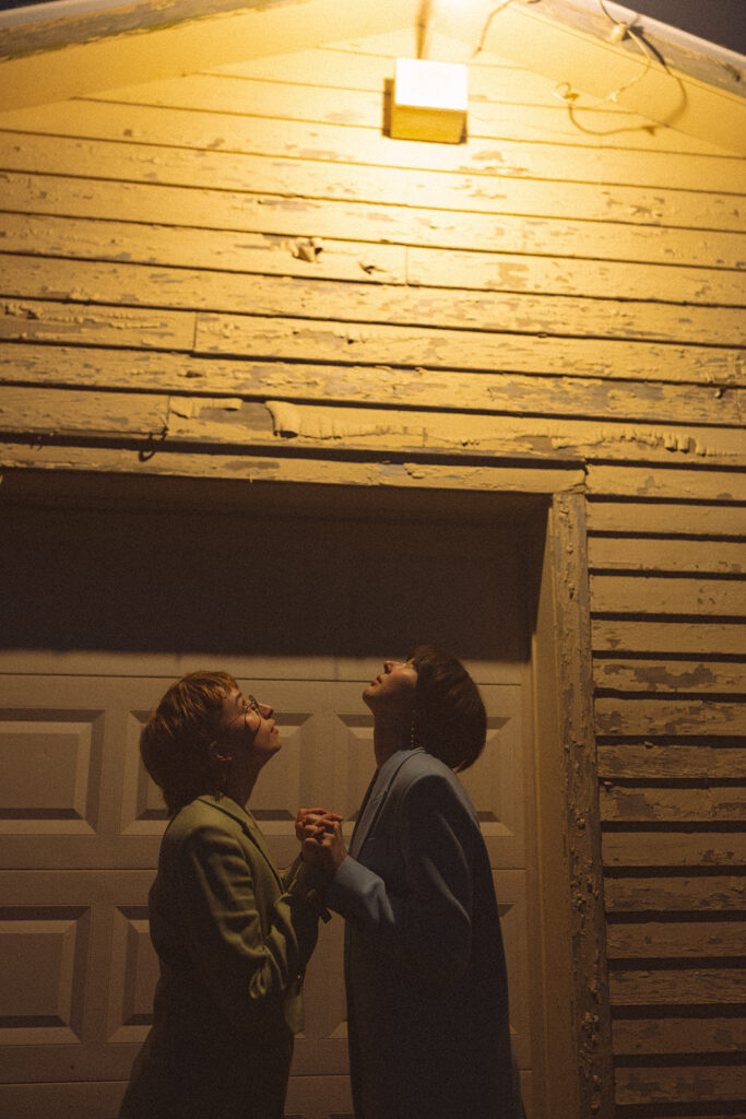 Couple holding hands beneath an outdoor light beside the weathered farmhouse garage for their unique Michigan engagement photos.