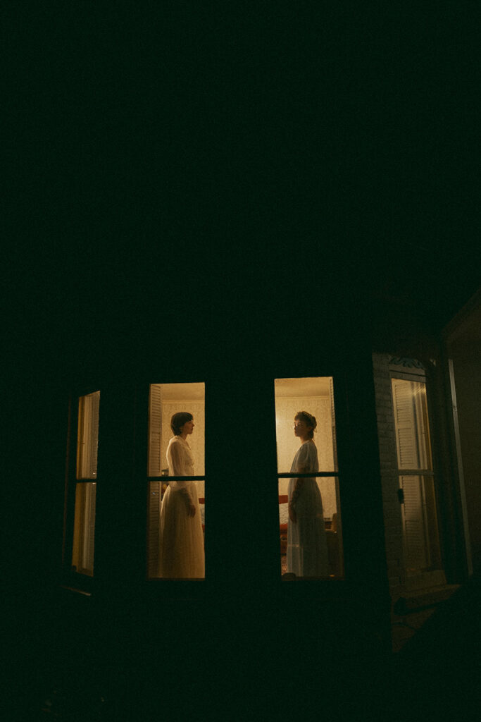 Couple facing each other through separate illuminated farmhouse windows at night.
