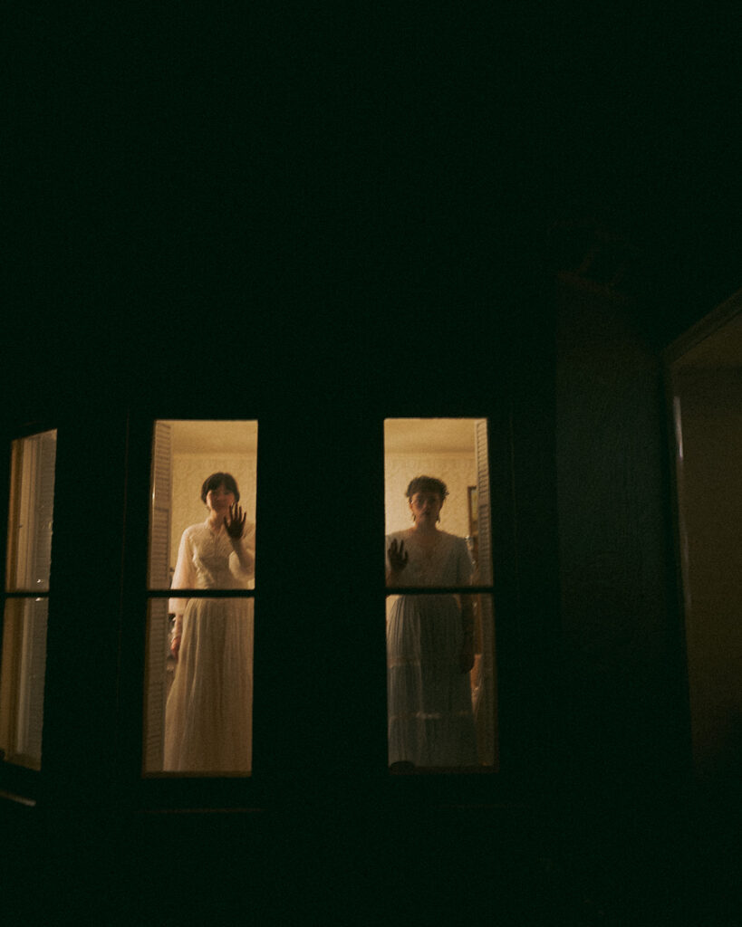 Couple raising their hands against the glowing farmhouse windows during their nighttime engagement photos for their unique Michigan engagement photos.