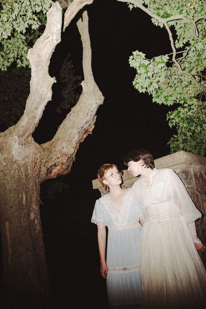 Couple in vintage dresses standing beneath a large tree at night with direct flash.