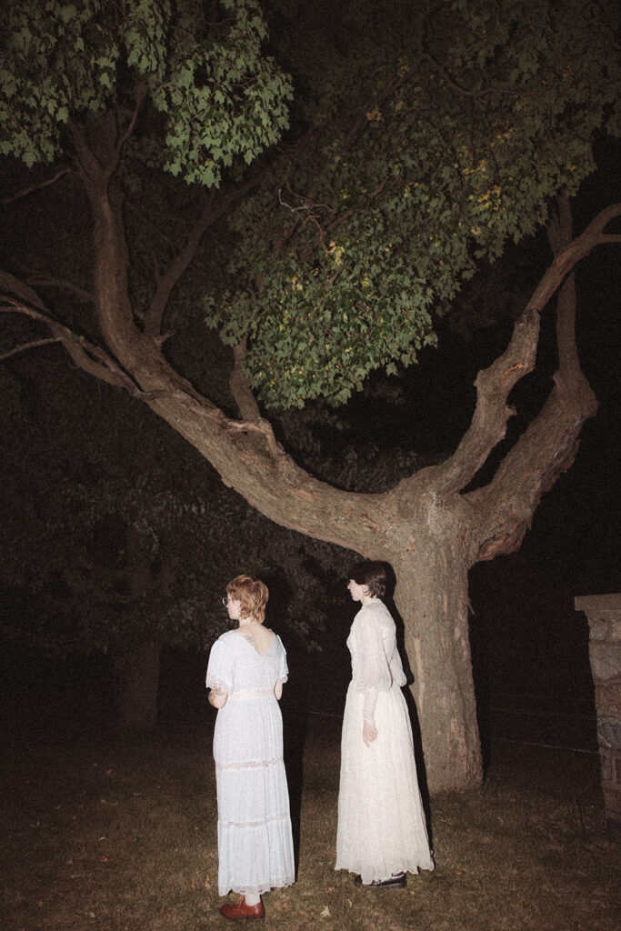 Couple in vintage dresses standing beneath a large tree at night with direct flash for for their unique Michigan engagement photos.