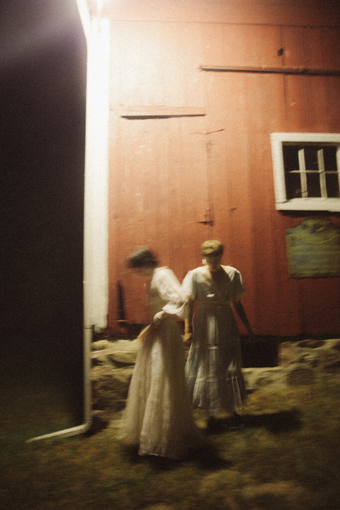Motion-blurred portrait of a couple in vintage dresses standing outside a red barn at night.