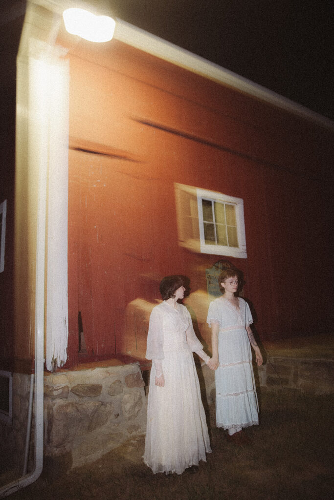 Motion-blurred portrait of a couple in vintage dresses standing outside a red barn at night.