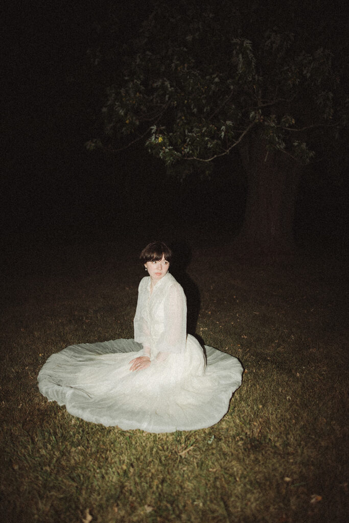 Person kneeling in the grass in a vintage dress at night.