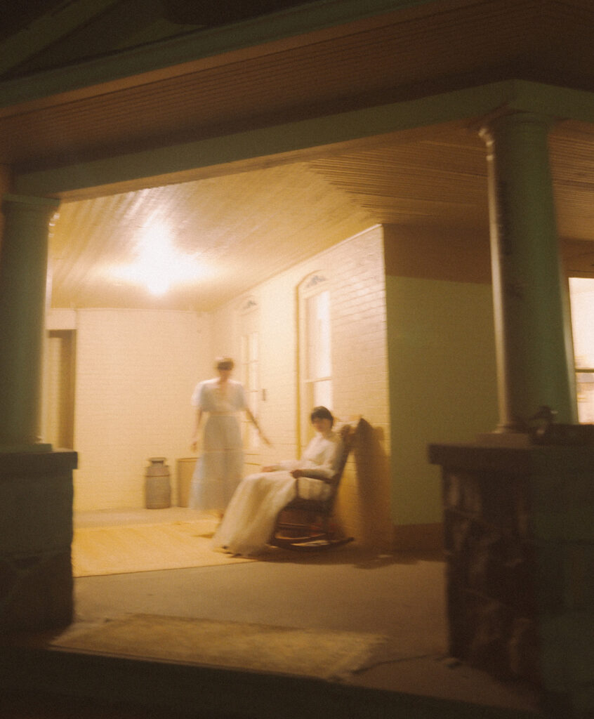 Couple in vintage dresses on the farmhouse porch, with one seated in a wooden rocking chair at night.