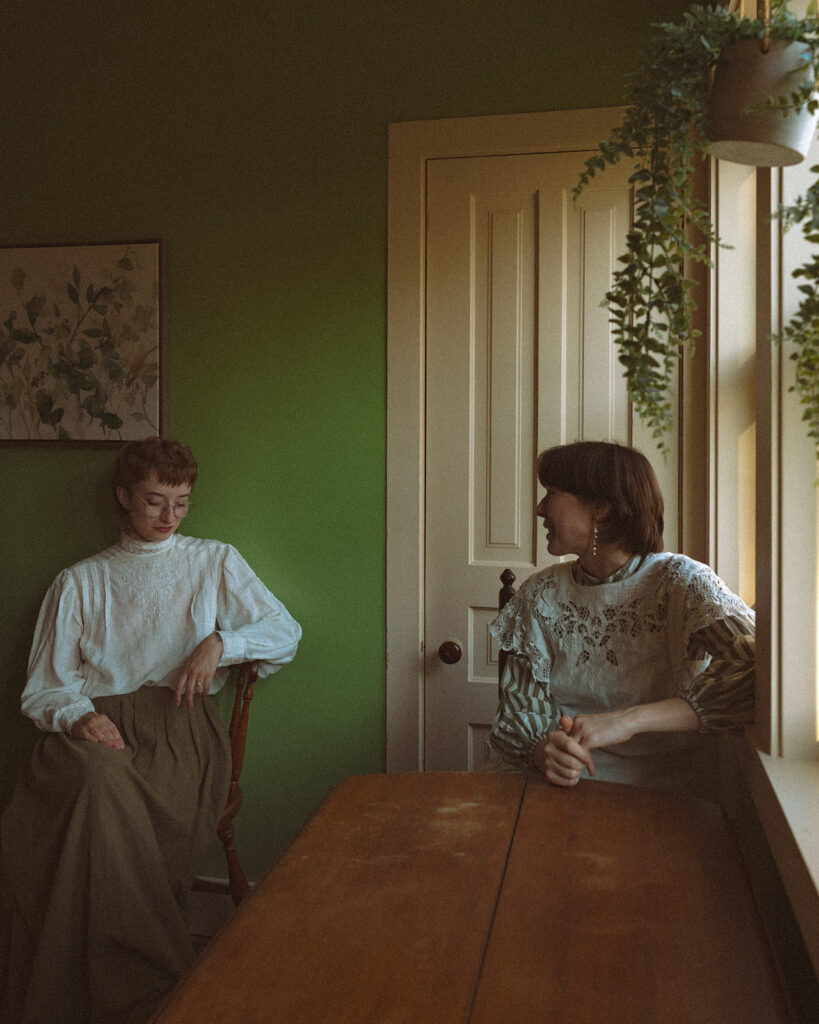 Couple in vintage-inspired clothing sitting beside a wooden table in a green farmhouse room.