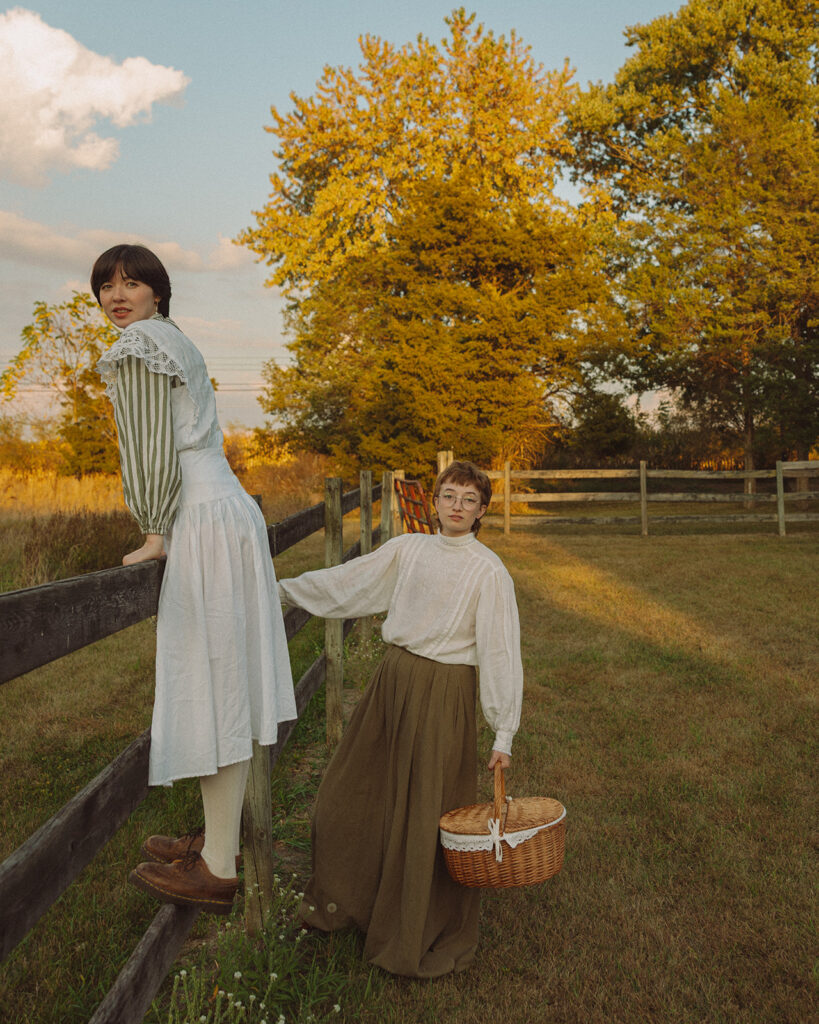 Couple in vintage-inspired clothing posing beside a wooden fence with a wicker picnic basket for their unique Michigan engagement photos.