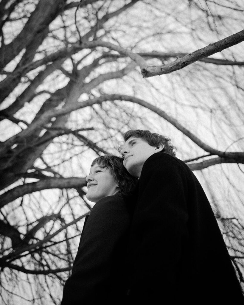 Black-and-white portrait of a couple standing close beneath bare tree branches.