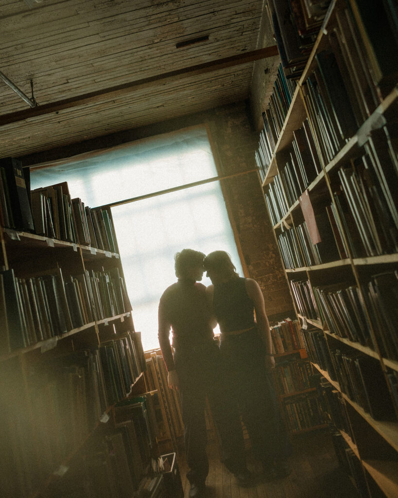 Moody silhouette of a couple standing close together at the end of a bookstore aisle.