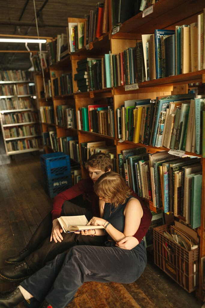Couple reading together on the floor beside colorful bookshelves during their indoor engagement session.