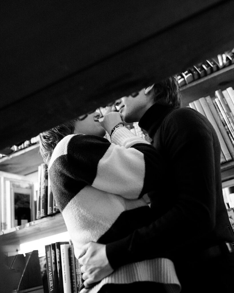 Black-and-white engagement photo of a couple playfully interacting between bookstore shelves.