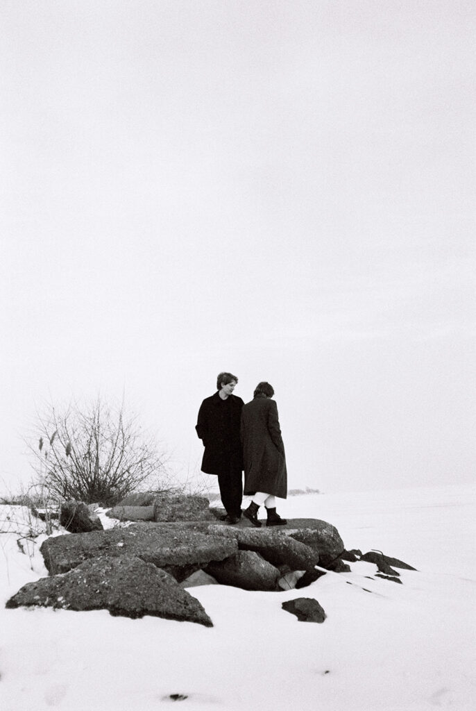 Black-and-white film photo of a couple standing on snowy rocks beside the frozen lake during their Detroit winter engagement session.