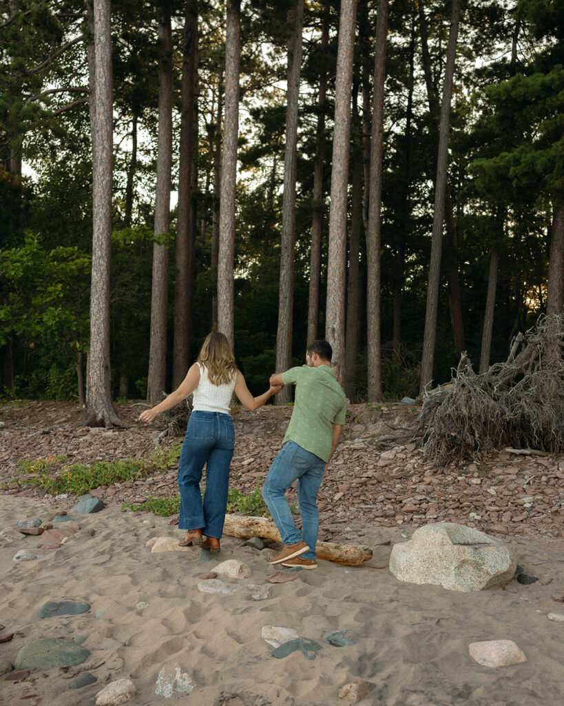 Couple holding hands beside driftwood and tall pine trees at Little Presque Isle.