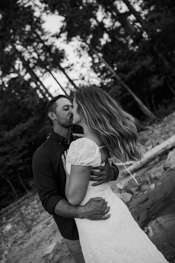 Black and white photo of a couple kissing with tall pines behind them.