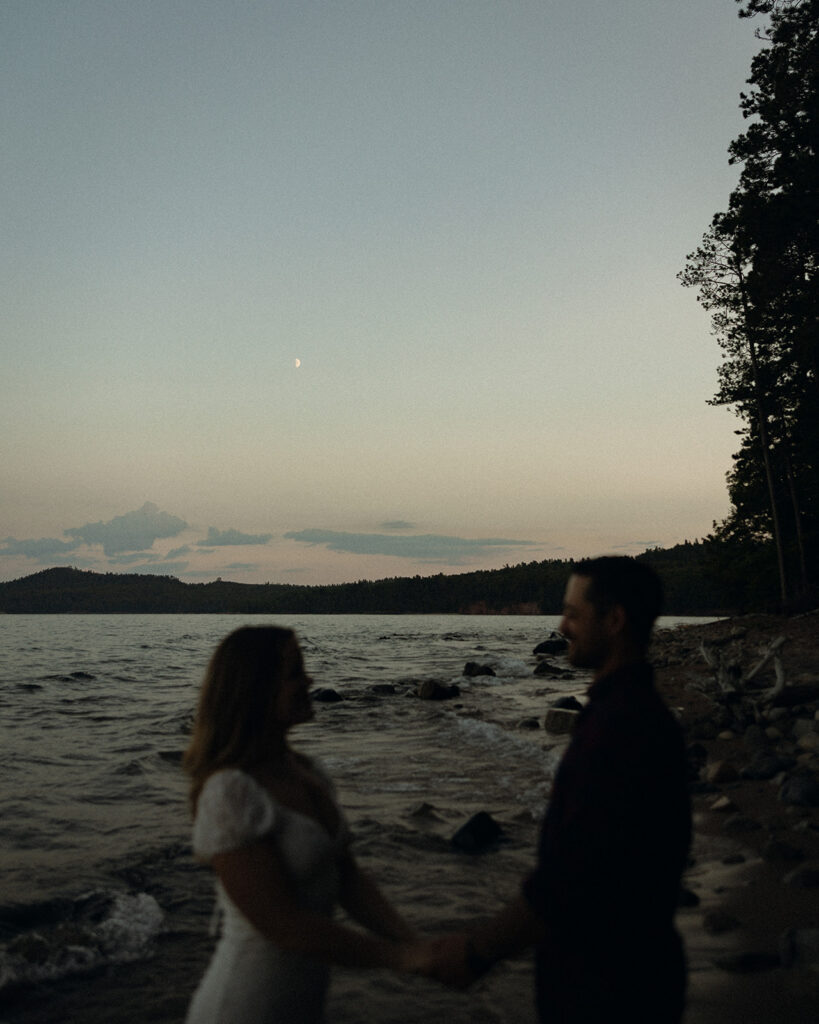 Silhouette of a couple holding hands beside Lake Superior beneath the moon at dusk.