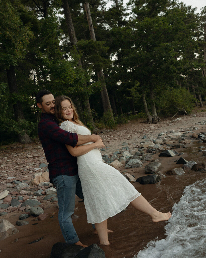 Man playfully lifting his fiancé along the shoreline of Lake Michigan.